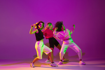 Flexible. Sportive girls dancing hip-hop in stylish clothes on purple-pink background at dance hall in green neon light. Youth culture, movement, style and fashion, action. Fashionable portrait.