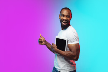 Wall Mural - Holding tablet, thumb up. African-american young man's portrait on gradient background in neon. Beautiful male model in casual style, white shirt. Concept of human emotions, facial expression, sales