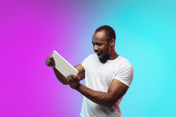 Wall Mural - Playing tablet, game. African-american young man's portrait on gradient background in neon. Beautiful male model in casual style, white shirt. Concept of human emotions, facial expression, sales, ad.