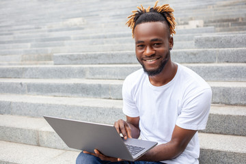 Freelance, technology, and education concept. Young smiling African American guy with lucky face working on laptop outdoors, sitting on the stairs in park, looking at camera. Empty copy space.