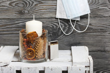 A Candle In A Glass Candlestick. The Candlestick Is Filled With Dried Cones, Spices And Dried Fruits. Stands On A Wooden Box. Medical Masks Hang Nearby.