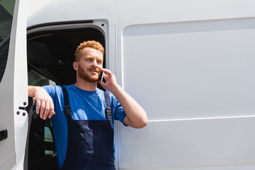 Loader in overalls talking on smartphone while standing near truck with open door outdoors