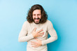 © Asier - Young long hair man isolated on a blue background laughs happily and has fun keeping hands on stomach.