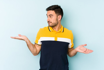 Young caucasian man isolated on blue background confused and doubtful shrugging shoulders to hold a copy space.