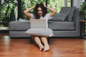 Furious and frustrated caucasian woman steaming and pulling her hair while sitting on the floor and working with laptop in living room.