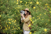 Smiling Slim Woman In Casual Wear And Hat Strolling With Old Video Camera And Leather Case Near Bushes With Colorful Blooming Flowers In Countryside And Looking Away