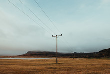 Electric Poles Located On Dry Grassy Terrain In Iceland Near Calm Sea Surrounded  By Rocky Mountains Against Foggy Sky