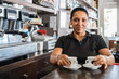 © Javier De La Torre/ADDICTIVE STOCK - Smiling ethnic female worker in black uniform standing at bar with cups of coffee at daytime