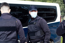 Anonymous Policeman In Sterile Mask And Uniform Standing Near Crop Unrecognizable Coworkers And Police Van During COVID 19 Pandemic At Work And Looking Away