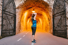 Side View Of Unrecognizable Fit Female Athlete In Sports Clothes And Armband Standing Near Rough Wall While Stretching Arms During Warm Up In Tunnel With Artificial Light And Metal Gates