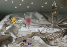 Above View Of Cocktail Decorated With Purple Flower Petals Served On White Tablecloth With Vintage Styled Glasses And Bottle On Sunny Day