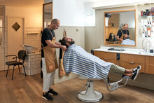 Barber Combing Hair While Cutting Hair To Serious Male Customer In Striped Cape In Modern Barbershop