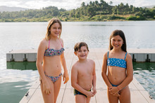 Smiling Brother And Sisters In Swimwear Standing On Quay Near Lake And Looking At Camera While Enjoying Summer Vacation