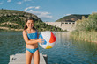 © Victor Gonzalez/ADDICTIVE STOCK - Happy little girl in bikini playing with beach ball standing in wooden pier near pond during summer holiday looking at camera