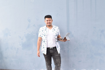 Indoor portrait of young man in t shirt standing against textured wall with copy space for ads, holding laptop and looking at camera with happy smile