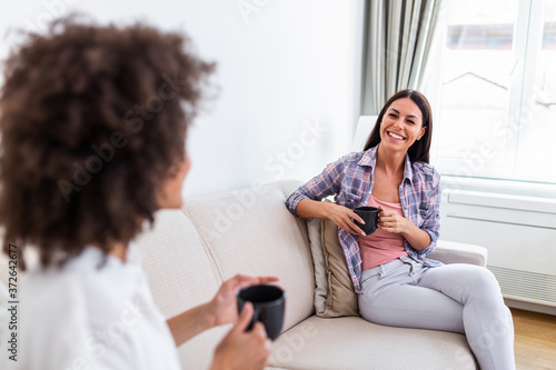 Two female friends in social distancing sitting on sofa. Best friends having coffee together while separated by social distancing on sofa at home