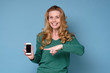 © Viktor Koldunov - Smiling mature blond hair woman holding mobile phone and pointing at it while standing against blue background