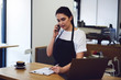 © BullRun - Attractive waitress talking on smartphone and making audit products at cafe indoors.