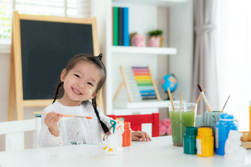 Asian kindergarten school girl painting Plaster doll with Acrylic water color paint and smiling looking at camera in living room at home. Homeschooling and distance learning..