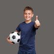 © Serhii - Handsome smiling fan boy holds soccer ball and gesturing thumbs up on gray background.