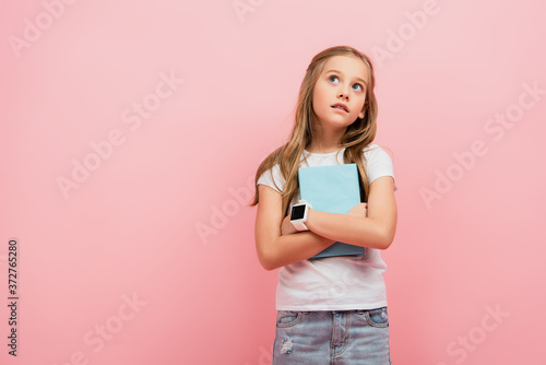 Fotografía thoughtful child in smartwatch looking away while holding book isolated on pink