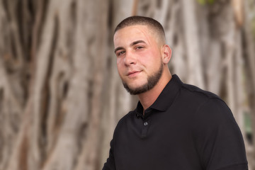 The handsome young man with a deep scar above his eye looks at the camera. A neatly trimmed chin hair matched by a military-style buzz hair cut in a black shirt compliments his rugged good looks.