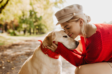 Wall Mural - Adorable girl and her dog sitting under green trees and bright sun in the park. Lovely blonde having good time together with her pet.