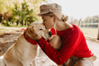 Beautiful blonde girl kissing her adorable dog in the autumn sunny park. Stylish young woman in red sweater and trendy hat holding tenderly the pet.