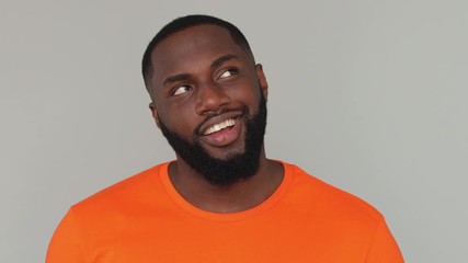 Wall Mural - Close up young dreamful bearded african american man guy 20s in orange basic casual t-shirt looking around think dream charming smile blinking wink eye isolated on grey color gray background in studio