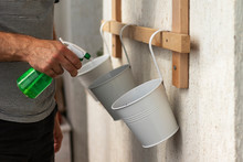 Man Sprays Pots With Planted Seeds. White Metal Pots For Plants Hang On Wall.