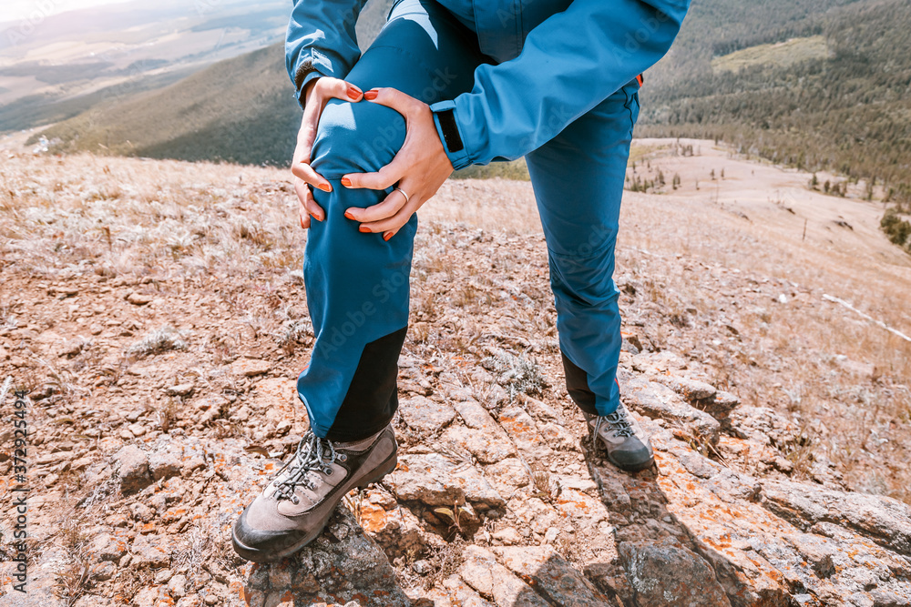 Woman is holding on to the knee joint, suffering from acute pain. Concept of injury of meniscus during a hike in the highlands - obrazy, fototapety, plakaty 