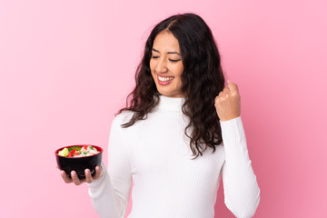 Canvas Print - Mixed race woman holding a bowl full of noodles over isolated pink background celebrating a victory