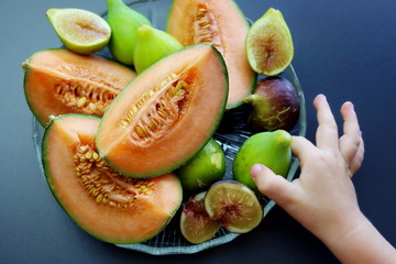 Wall Mural - Slices of ripe and juicy melon and sweet figs in a round plate on a gray background. A child's hand reaches for the fruit. A plate with autumn fruits.