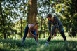 © Sanja - Young couple doing a muscle warming exercise in nature, woman smiles while man looks at her