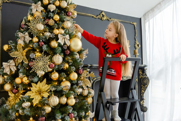 little blonde girl in a red sweater decorates a christmas tree.