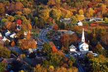 A Classic New England Town, With A White Church With A Large Steeple, Is Surrounded By Brilliant Autumn Foliage In An Aerial View