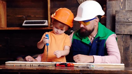 Wall Mural - Father and his little son with tools in workshop. Boy child busy in helmet learning to use hammer with dad. Early childhood education concept.