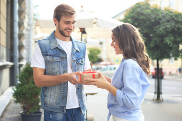 Wall Mural - Astonished excited couple in summer clothes smiling and holding present box together while standing on city street.