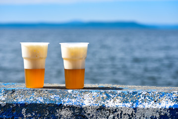 Two disposable glasses of beer on an embankment parapet against blue sea background