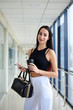 © Natalia - Young brunette woman, wearing white pants and black top, holding black purse, cell phone and paper coffee cup, standing in light office building. Businesswoman on lunch break.