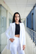© Natalia - Young brunette woman, wearing white pants and jacket, standing in light passageway with huge windows, posing for social media. Businesswoman on a break. Female portrait photography.