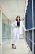 © Natalia - Young brunette woman, wearing white pants and jacket, standing in light passageway with huge windows, posing for social media, leaning on railing. Businesswoman on a break. Female portrait photography