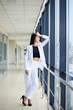 © Natalia - Young brunette woman, wearing white pants and jacket, standing in light passageway, posing for social media, leaning on window. Businesswoman on a break. Female portrait photography.