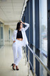© Natalia - Young brunette woman, wearing white pants and jacket, standing in light passageway, posing for social media, leaning on window. Businesswoman on a break. Female portrait photography.