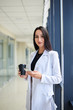 © Natalia - Young brunette woman, wearing white business suit, holding cell phone and black paper coffee cup, standing in light office building near window. Businesswoman on lunch break. Corporate culture concept