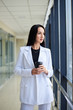 © Natalia - Young brunette woman, wearing white business suit, holding black paper coffee cup, standing in light office building near window. Businesswoman on lunch break. Corporate culture concept.