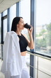 © Natalia - Young brunette woman, wearing white business with jacket in shoulder, drinking coffee from black paper to go cup, standing in light office building near window. Businesswoman on lunch break.