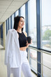 © Natalia - Young brunette woman, wearing white business with jacket in shoulder, holding black paper coffee cup, standing in light office building near window. Businesswoman on lunch break. Corporate culture.