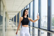 © Natalia - Young brunette woman, wearing white pants and black top, holding jacket in hand, taking selfie picture with cell phone in light passageway, posing for social media. Businesswoman on lunch break.