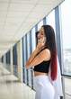 © Natalia - Young brunette woman, wearing white pants and black top, holding jacket in hand, talking on cell phone in light passageway of office building. Businesswoman on lunch break. Corporate culture concept.